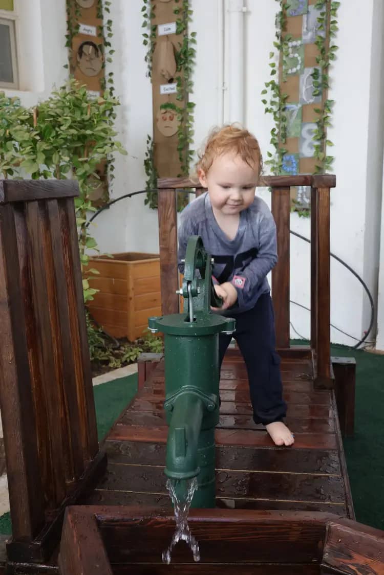 Young child enjoying play while operating a green hand water pump on a wooden platform, with water flowing into a basin and educational wall decorations in the background.