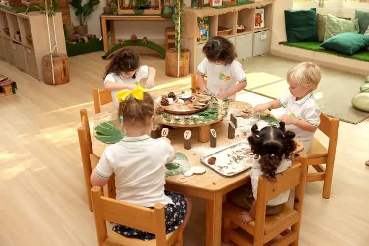 Young child in a green shirt with pink design playing at a table outdoors with jars, teapot, and containers in a courtyard setting.