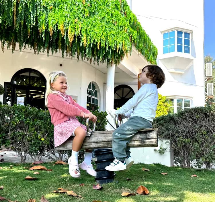 Two children sitting on a wooden seesaw in a garden area with green grass, dry leaves, and a white building with arched windows and hanging plants in the background.