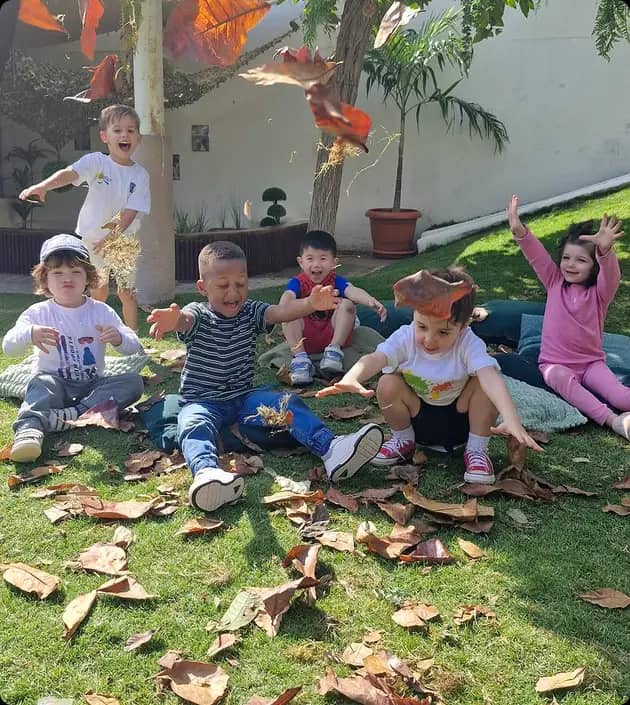 Group of young children sitting on grass and joyfully tossing dry autumn leaves in the air in a garden setting with trees and plants in the background.